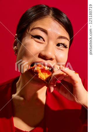Close-up of satisficed Asian woman biting spicy crispy chicken nuggets with tomato sauce from carton box to go against red background. 125180719