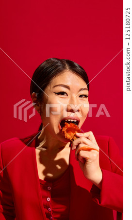 Close-up portrait of Asian woman in red eating spicy chicken nuggets with tomato sauce on top against bold red background. Close-up portrait of Asian woman in red eating spicy chicken nuggets with tomato sauce on top against bold red background. 125180725