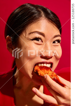 Close-up portrait of young excited Asian woman biting crispy chicken dripping with sauce against bold red background. 125180735