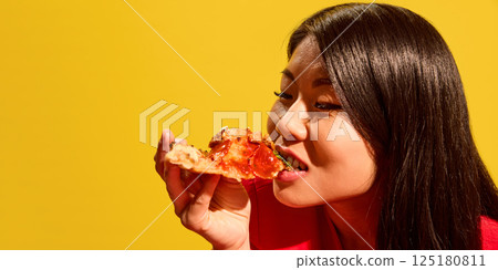 Close side profile of young Asian woman joyfully eating pizza slice with tomato sauce against bright yellow background. Close side profile of young Asian woman joyfully eating pizza slice with tomato sauce against bright yellow background. 125180811