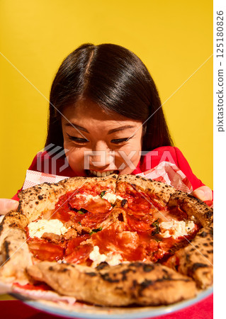 Woman in red bites giant pizza slice with intense hunger and playful face against bright yellow background. Delicious Italian cuisine. 125180826