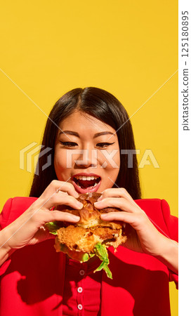 Excited Asian young woman in red prepares to bite large sandwich while looking at it with open mouth and wide smile against bright yellow background. Excited Asian young woman in red prepares to bite large sandwich while looking at it with open mouth and wide smile against bright yellow background. 125180895