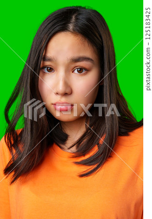 Teen girl with long dark hair looks thoughtfully ahead against a bright green background wearing an orange shirt Teen girl with long dark hair looks thoughtfully ahead against a bright green background wearing an orange shirt 125183425