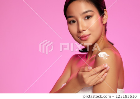 Young woman applying skin cream against a vibrant pink background during a beauty routine session Young woman applying skin cream against a vibrant pink background during a beauty routine session 125183610