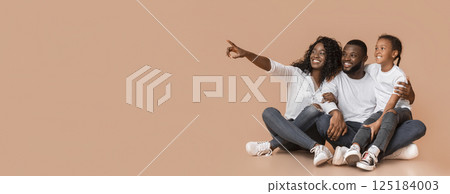 Happy black family of three sitting on floor and pointing aside at copy space over yellow background, panorama 125184003