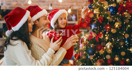 Happy family of three in red Santa hats decorating xmas tree with bubbles and lights at home 125184004