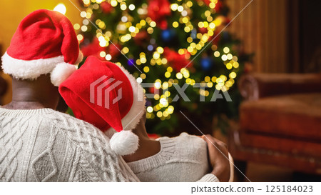 Romantic afro couple sitting next to Christmas tree, wearing Santa's hats and hugging, rear view 125184023
