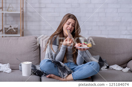 Comfort foods and stress eating concept. Frustrated young lady devouring pastry on sofa at home. Upset woman dealing with negative emotions, anxiety or problems through sweets Comfort foods and stress eating concept. Frustrated young lady devouring pastry on sofa at home. Upset woman dealing with negative emotions, anxiety or problems through sweets 125184074
