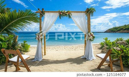 A picturesque wedding altar is set on the sandy beach, adorned with flowers and draped fabric, overlooking calm turquoise waters and a bright blue sky. 125184077