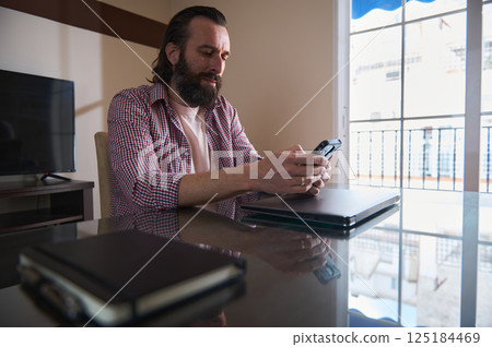 Bearded Man Typing on Phone at Home Office Desk Near Large Window Bearded Man Typing on Phone at Home Office Desk Near Large Window 125184469