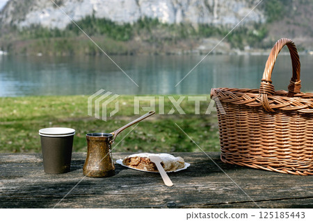 Rustic picnic setup by mountain lake. Woven basket coffee pot cup and wrapped pastry Austrian strudel on wooden table. Serene nature backdrop with calm water green grass and mountains. 125185443