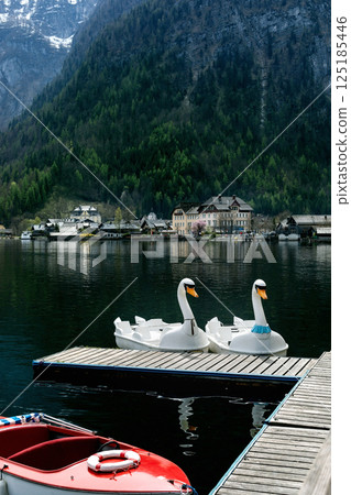 Hallstatt Austria. Charming lakeside village nestled among mountains with swanshaped pedal boats docked at wooden pier. Red rescue boat visible. Alpine forest backdrop creates peaceful atmosphere. Hallstatt Austria. Charming lakeside village nestled among mountains with swanshaped pedal boats docked at wooden pier. Red rescue boat visible. Alpine forest backdrop creates peaceful atmosphere. 125185446