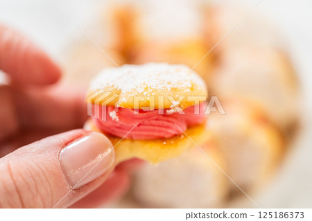 A bowl filled with lemon spritz cookie sandwiches, each piped with vibrant cranberry buttercream and dusted with powdered sugar. The cookies have a delicate, buttery texture with a festive touch 125186373
