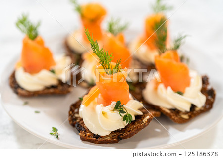 A gourmet smoked salmon canape elegantly arranged on a crispy cracker, topped with a swirl of smooth cream cheese, fresh thyme, and a drizzle of olive oil. The vibrant orange salmon contrasts A gourmet smoked salmon canape elegantly arranged on a crispy cracker, topped with a swirl of smooth cream cheese, fresh thyme, and a drizzle of olive oil. The vibrant orange salmon contrasts 125186378