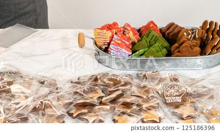 Captured from a mid-range perspective, a person carefully packages a decorated Christmas tree-shaped cookie while an assortment of festive treats, including green, red, and gingerbread cookies, is 125186396