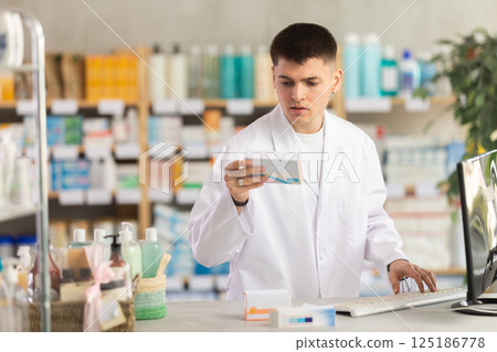 Professional young man pharmacist standing at prescription counter and working on computer, stocktaking medicines at pharmacy shop 125186778