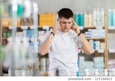 Sick man standing between shelves in drugstore suffering from headache or having migraine, choosing medicine. Health care and medicine concept 125186822