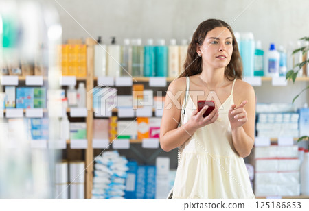 Woman is standing in a pharmacy and looking for goods with a phone in her hands Woman is standing in a pharmacy and looking for goods with a phone in her hands 125186853