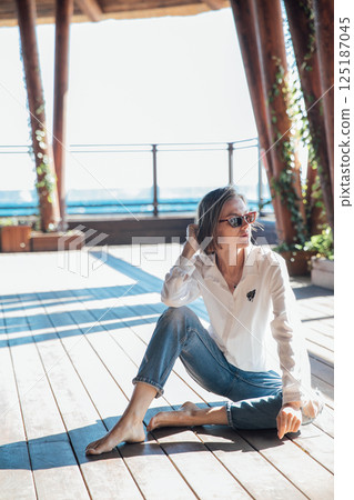 woman in sunglasses sitting on wooden floor on beach by sea 125187045