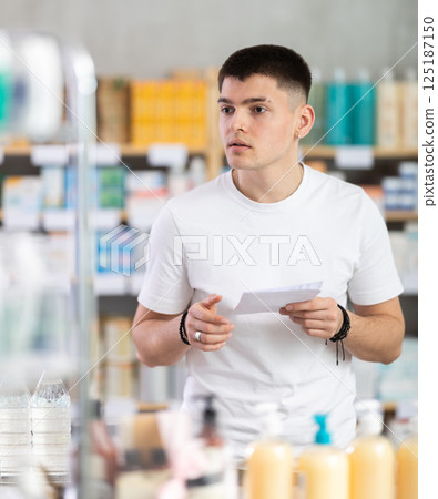 Portrait of young guy with prescription in hand looking for something in drugstore 125187150
