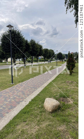 paved road in european village surrounded by neatly planted trees, trimmed grass and blue sky, pink and white bricks go well with blue clouds paved road in european village surrounded by neatly planted trees, trimmed grass and blue sky, pink and white bricks go well with blue clouds 125187461