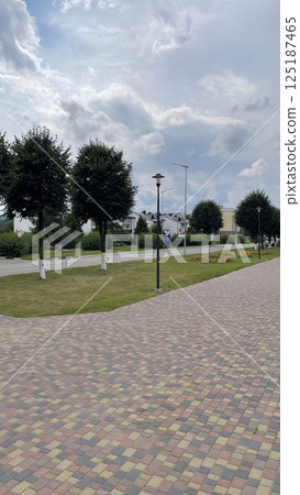 paved road in european village surrounded by neatly planted trees, trimmed grass and blue sky, pink and white bricks go well with blue clouds paved road in european village surrounded by neatly planted trees, trimmed grass and blue sky, pink and white bricks go well with blue clouds 125187465
