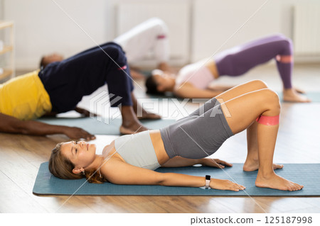 Girl performs glute bridge during Pilates multinational training with elastic band on shins Girl performs glute bridge during Pilates multinational training with elastic band on shins 125187998