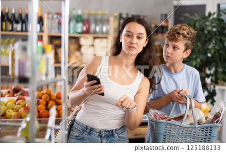 Mom chooses products from a list on her smartphone. Teenage son helps carry shopping cart in supermarket 125188133