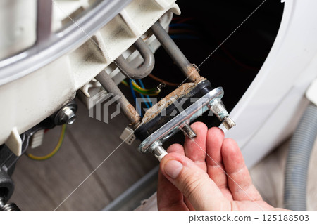 Electric heating element in hand against the background of a washing machine. Damage to the heating element in the washing machine due to hard water, close-up 125188503