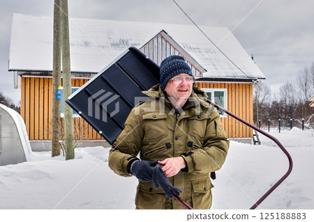 Carrying wide snow shovel over shoulder mature white man in warm coat and knitted hat appears focused while standing near wooden house in snowy setting. Carrying wide snow shovel over shoulder mature white man in warm coat and knitted hat appears focused while standing near wooden house in snowy setting. 125188883
