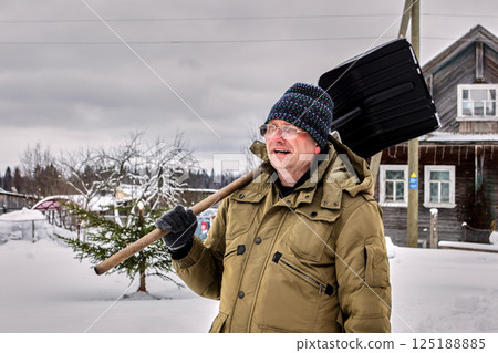 Person wearing blue knit hat and olive green winter jacket carries snow shovel over shoulder while standing in snowy rural area with wooden house and bare trees in background. Person wearing blue knit hat and olive green winter jacket carries snow shovel over shoulder while standing in snowy rural area with wooden house and bare trees in background. 125188885