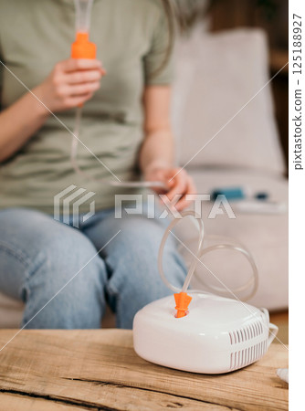 Woman using a nebulizer for respiratory treatment at home, sitting on a couch with medical equipment on the table. 125188927