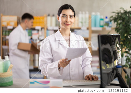 Girl pharmacist with prescription uses computer at workplace. Colleague work in background Girl pharmacist with prescription uses computer at workplace. Colleague work in background 125191182