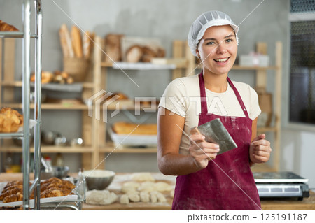 Dedicated young female baker standing with dough scraper in hands Dedicated young female baker standing with dough scraper in hands 125191187