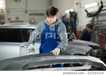 Male mechanic prepares car for painting in workshop - using grinding machine he sands bumper before painting 125191229