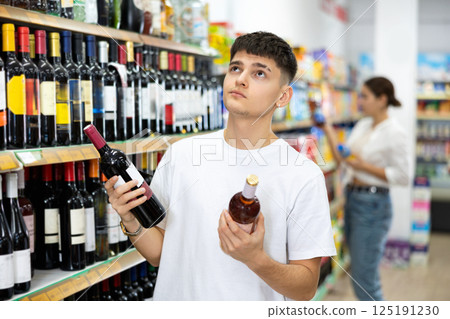 in supermarket young man carefully chooses wine in supermarket young man carefully chooses wine 125191230