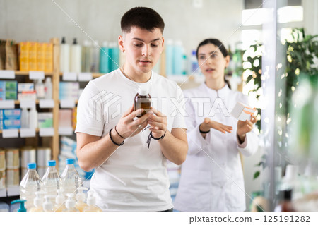Guy chooses cough syrup in modern pharmacy. Female pharmacist helps with choosing medicine Guy chooses cough syrup in modern pharmacy. Female pharmacist helps with choosing medicine 125191282