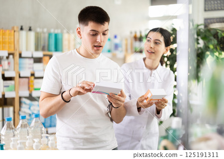 Guy chooses pills in modern pharmacy. Female pharmacist helps with choosing medicine Guy chooses pills in modern pharmacy. Female pharmacist helps with choosing medicine 125191311