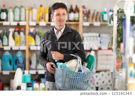 Positive man selects various household cleaning products for cleaning the house with full basket of various purchases Positive man selects various household cleaning products for cleaning the house with full basket of various purchases 125191343