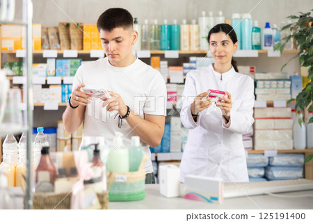 Guy chooses pills paracetamol in modern pharmacy. Female pharmacist helps with choosing medicine 125191400