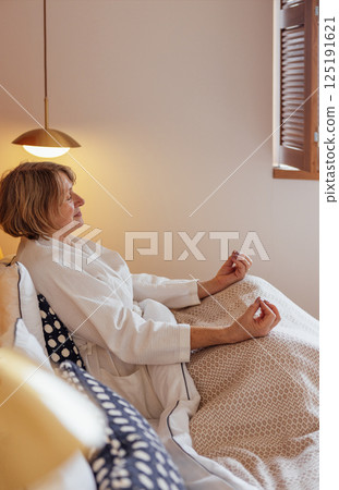 An elderly woman in a white coat is sitting in bed and meditating 125191621
