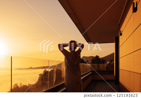 Young woman in a dress is enjoying the sunrise. The lady stands on the balcony and looks at the morning misty landscape. 125191623