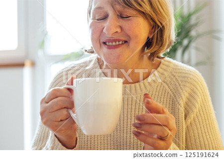 Close-up of an elderly woman in a knitted sweater drinking her morning coffee in the kitchen. A beautiful middle-aged lady in casual clothes is enjoying tea at home. 125191624
