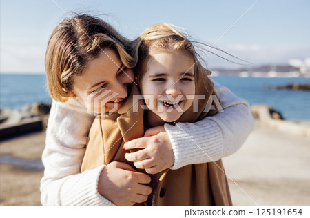 A smiling mother hugs her cute laughing daughter on the seashore. A young woman puts on a warm coat for her child on the beach. 125191654