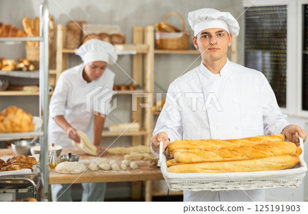 Young male baker holding baguettes in wicker basket Young male baker holding baguettes in wicker basket 125191903
