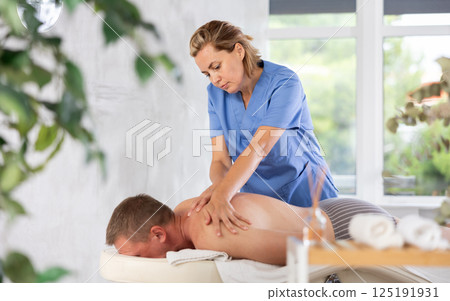 Female masseur performs back stroking and patting during massage session for male client Female masseur performs back stroking and patting during massage session for male client 125191931