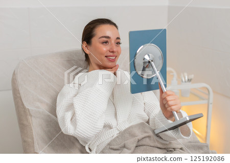 Caucasian young woman smiles in mirror after facial therapy in beauty clinic showing visible result Caucasian young woman smiles in mirror after facial therapy in beauty clinic showing visible result 125192086