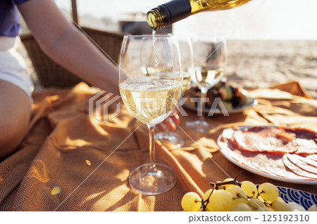 Close-up of a glass of champagne on a picnic mat. The cup is filled with sparkling wine outdoors on the beach. Delicious snacks and drinks by the sea. 125192310