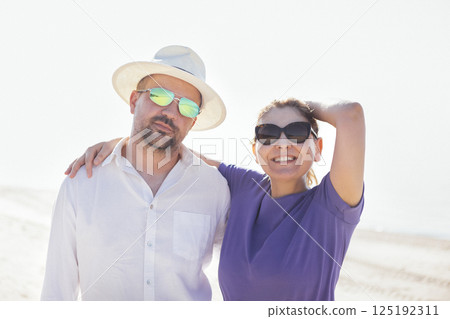 A young married couple in casual clothes is laughing by the sea. An attractive woman in sunglasses smiles and hugs her husband on the beach. 125192311
