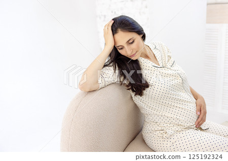A young thoughtful pregnant woman is sitting on an armchair. An attractive expectant mother gently touches her stomach at home. A young thoughtful pregnant woman is sitting on an armchair. An attractive expectant mother gently touches her stomach at home. 125192324